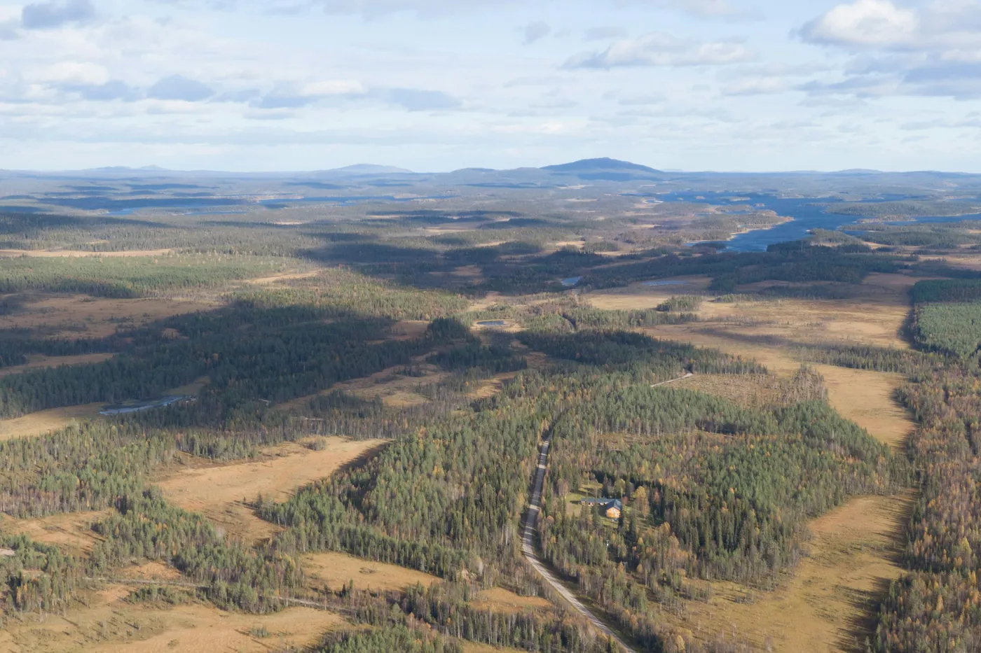 Aerial view of a wide forested landscape with bogs, a lake and a gravel road, low fells on the horizon under a partly cloudy sky