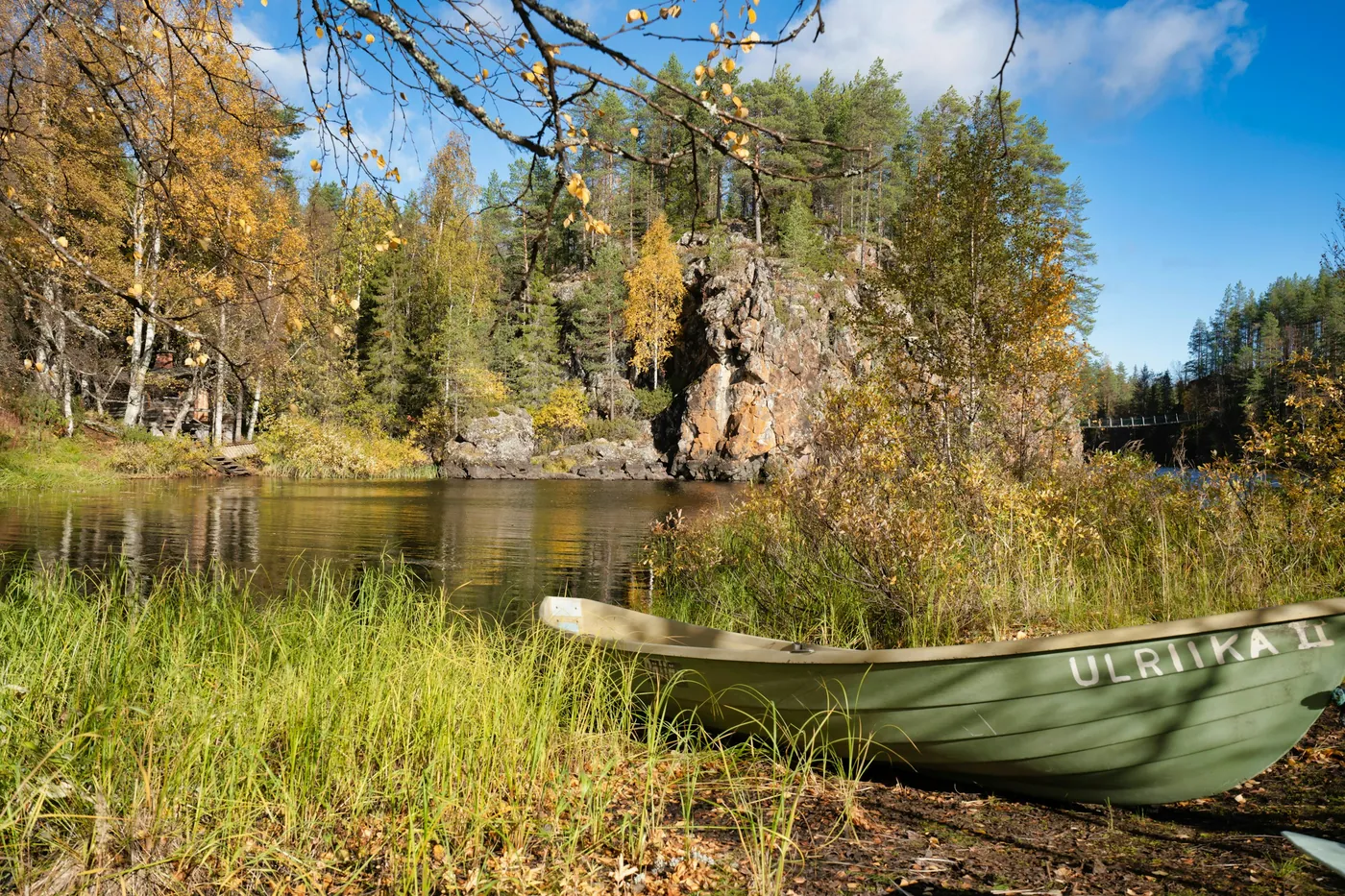 Une barque en bois verte tirée sur une berge herbeuse, avec une eau calme, une paroi rocheuse rougeâtre et des bouleaux aux feuilles jaunes derrière