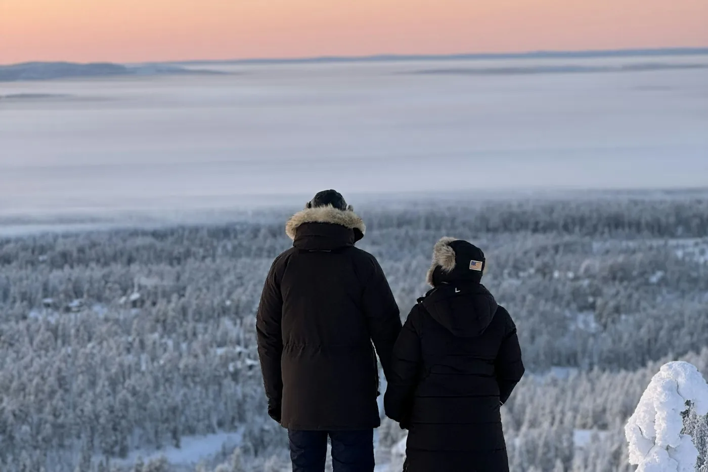 Two people in dark winter parkas with fur-trimmed hoods seen from behind, looking out over a snowy forest under a pink evening sky