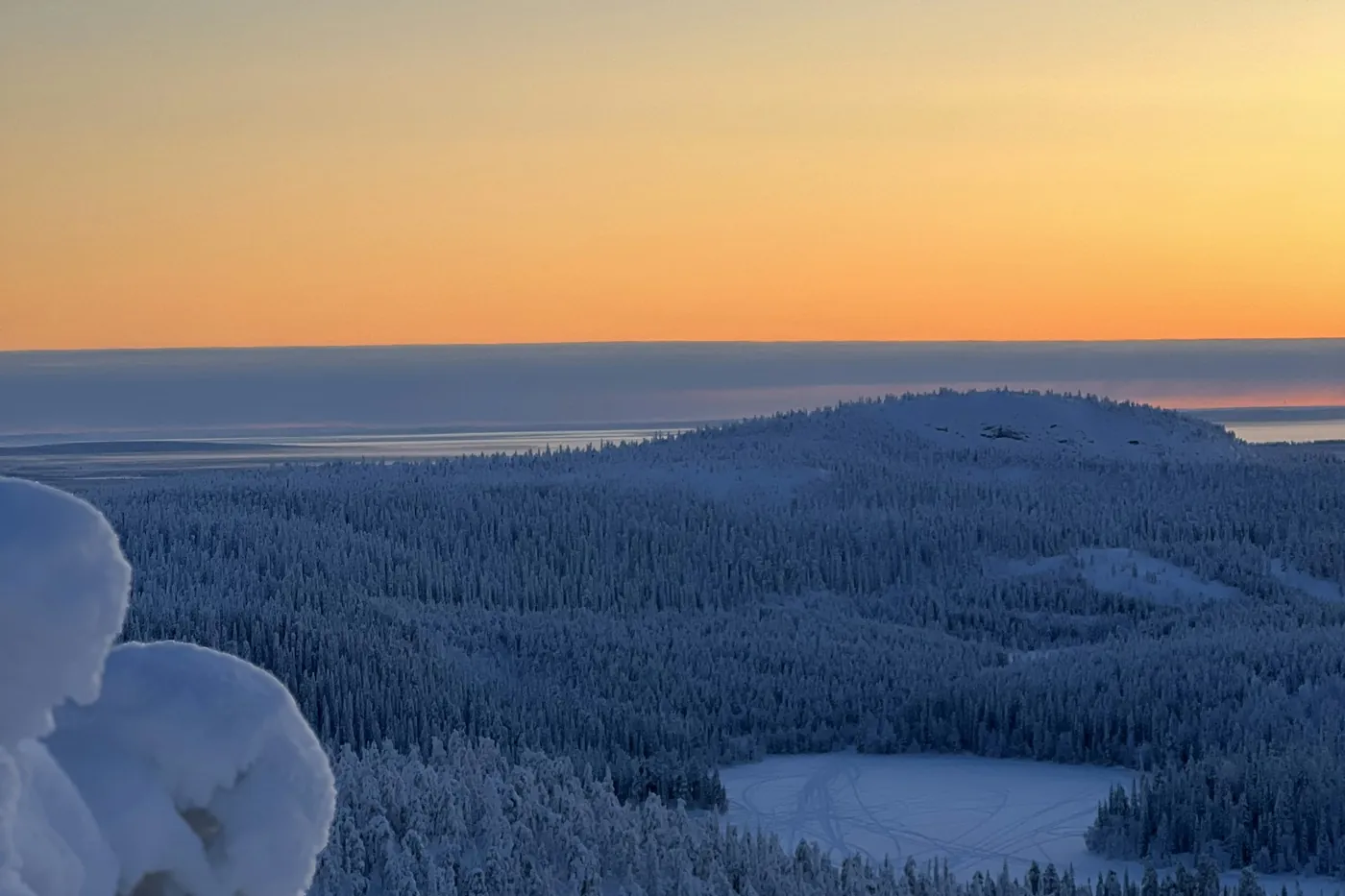 An orange and yellow sky above a band of low cloud, with a snow-covered forested fell and a frozen pond in blue shadow below