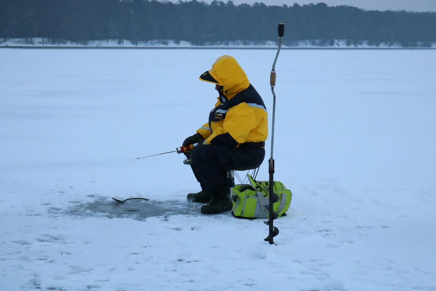 A person in a yellow and black winter suit sits on a stool on a frozen lake, fishing through a hole in the ice, an ice drill standing beside them