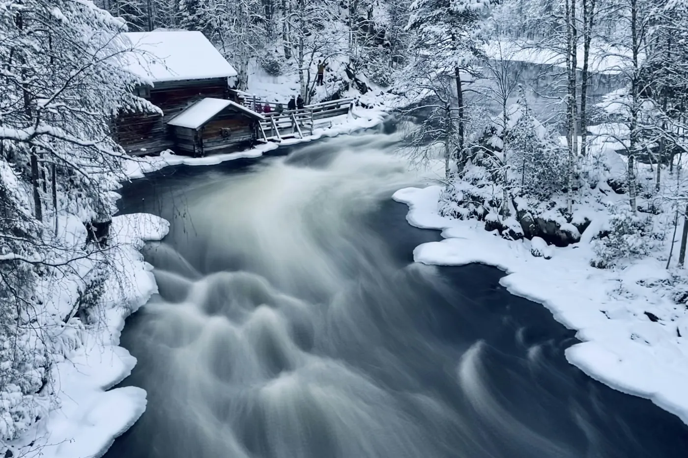 A fast river with blurred white water running past an old wooden mill building and a smaller hut, all covered in snow, with a footbridge and a few people behind
