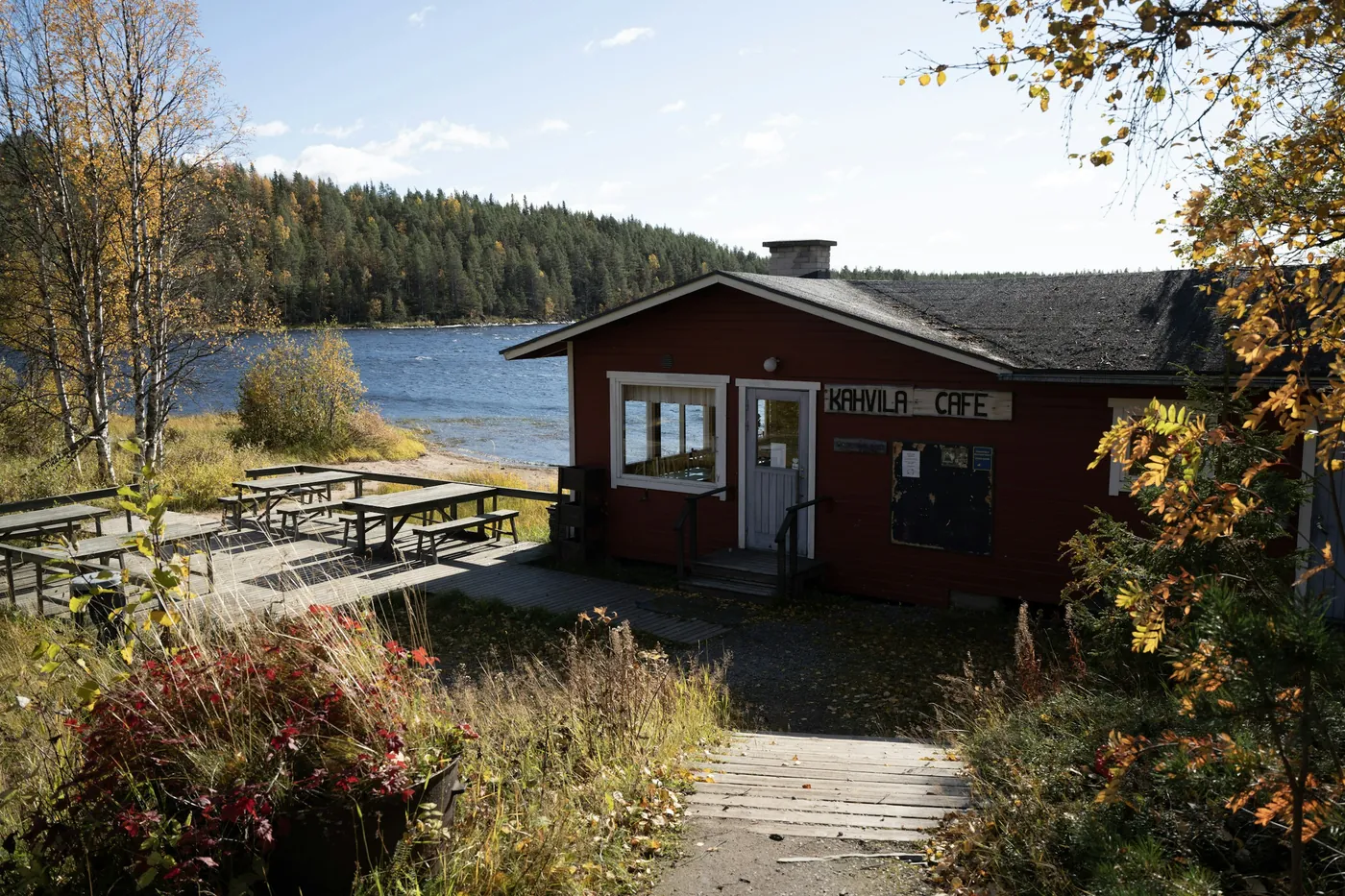 A red wooden building with a sign reading Kahvila Cafe beside a lake, picnic tables on a paved terrace and birch trees in yellow autumn leaves