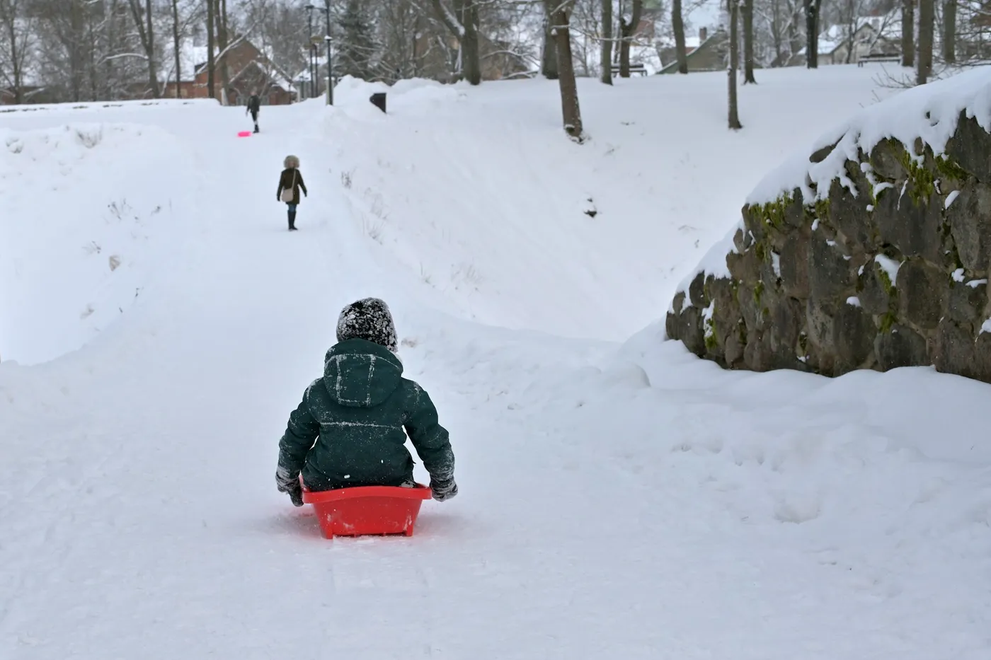 A small child in a dark green snowsuit and knitted hat sliding down a snowy path on a red plastic sledge, two figures walking further up the hill